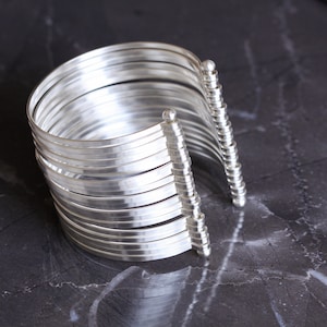 May include: A silver cuff bracelet with multiple parallel bands. The bracelet has a curved design and is open on one side, with decorative vertical posts on each end. The jewelry is displayed on a dark, marbled surface.