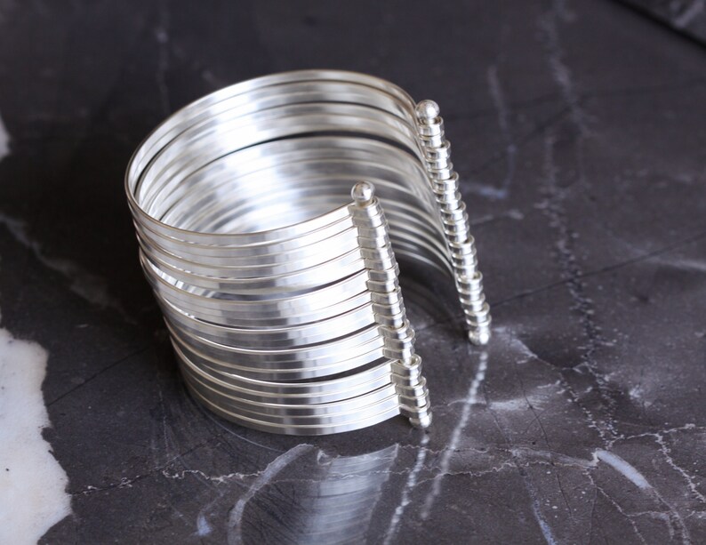 May include: A silver cuff bracelet with multiple parallel bands. The bracelet has a curved design and is open on one side, with decorative vertical posts on each end. The jewelry is displayed on a dark, marbled surface.
