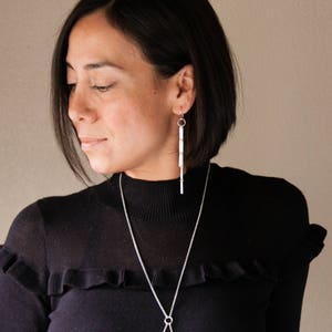Peut inclure: Une personne portant un pull &agrave; col roul&eacute; noir avec des boucles d'oreilles argent&eacute;es et un collier argent&eacute;. Les boucles d'oreilles sont de longues barres rectangulaires. Le collier a une longue cha&icirc;ne avec un pendentif circulaire.