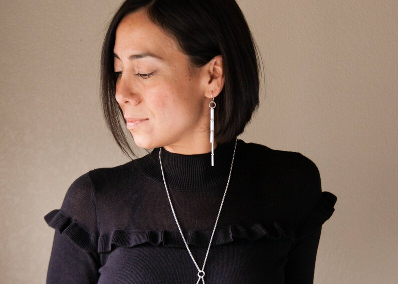 Peut inclure: Une personne portant un pull &agrave; col roul&eacute; noir avec des boucles d'oreilles argent&eacute;es et un collier argent&eacute;. Les boucles d'oreilles sont de longues barres rectangulaires. Le collier a une longue cha&icirc;ne avec un pendentif circulaire.