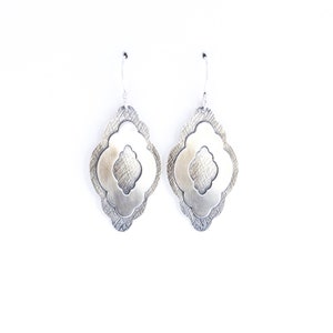 Pode incluir: Um par de brincos pendurados prateados. Cada brinco apresenta um design em camadas com uma camada externa texturizada e uma camada interna lisa vazada. Os brincos penduram-se em ganchos de prata simples, sobre um fundo branco.