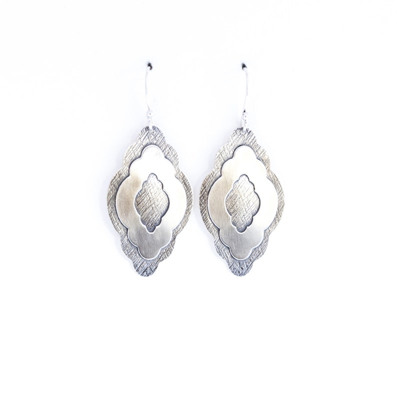 Pode incluir: Um par de brincos pendurados prateados. Cada brinco apresenta um design em camadas com uma camada externa texturizada e uma camada interna lisa vazada. Os brincos penduram-se em ganchos de prata simples, sobre um fundo branco.