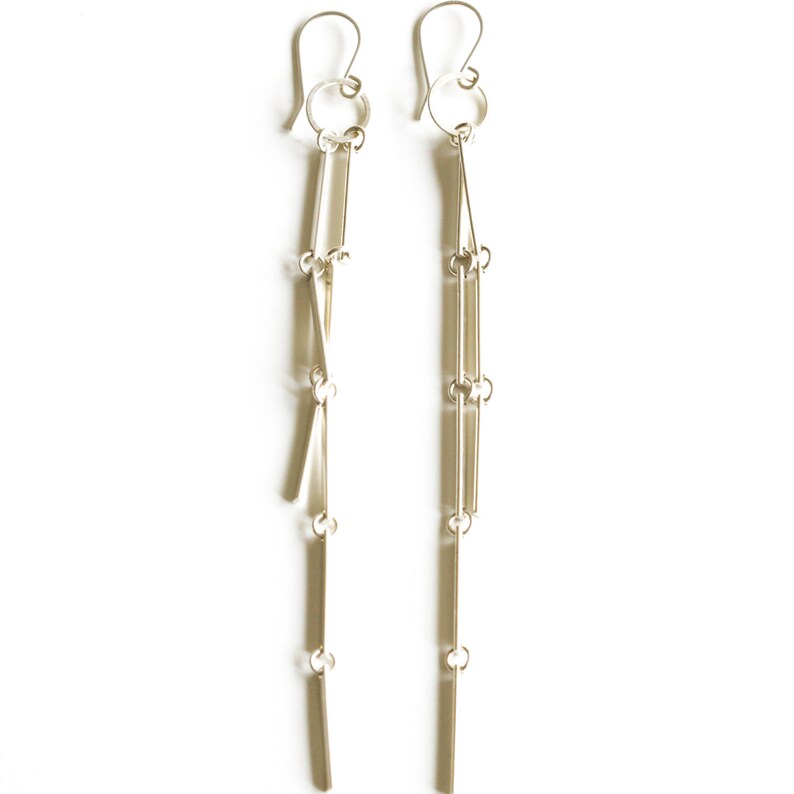 Peut inclure: Une paire de boucles d'oreilles pendantes argent&eacute;es. Chaque boucle d'oreille comprend un crochet, un petit anneau et plusieurs barres rectangulaires verticales reli&eacute;es par de petits anneaux, cr&eacute;ant un design lin&eacute;aire et fluide. Sur fond blanc.