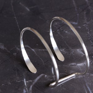 Puede incluir: Una pulsera de pu&ntilde;o plateada con un dise&ntilde;o minimalista. La pulsera presenta dos bandas curvas y abiertas conectadas por una barra recta. El metal tiene una superficie pulida y reflectante, y la forma general es suave y moderna.