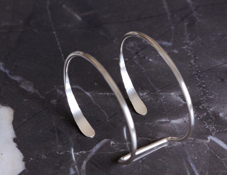 Puede incluir: Una pulsera de pu&ntilde;o plateada con un dise&ntilde;o minimalista. La pulsera presenta dos bandas curvas y abiertas conectadas por una barra recta. El metal tiene una superficie pulida y reflectante, y la forma general es suave y moderna.