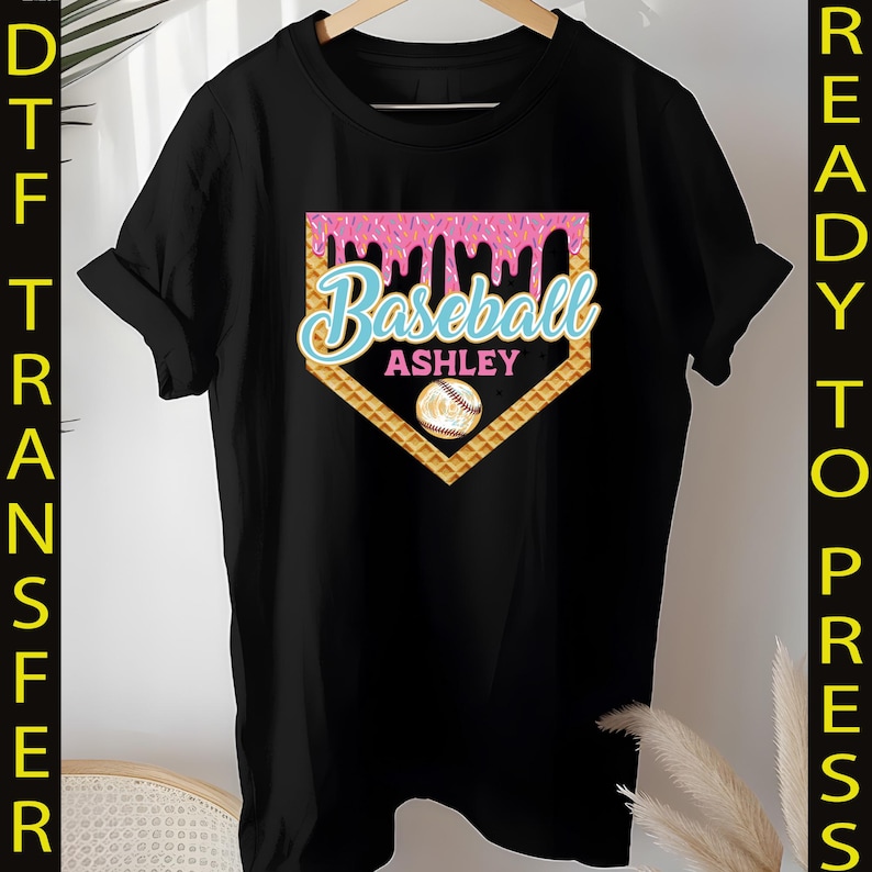 May include: Black t-shirt with a pink and yellow baseball graphic. The graphic features a waffle cone design with pink frosting and sprinkles. The text "Baseball Ashley" is written in blue on the graphic.
