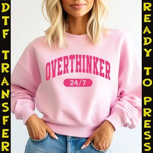 May include: A light pink sweatshirt with the word "OVERTHINKER" in bold, pink letters across the chest, with "24/7" below. The sweatshirt is a crew neck style.