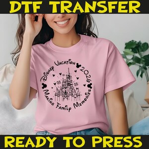 K&ouml;nnte beinhalten: Hellrosa T-Shirt mit schwarzer Grafik eines Schlosses, Mickey Mouse-Silhouetten und den Worten "Disney Vacation 2026 Making Family Memories". Das Shirt hat auch den Text "DTF TRANSFER" und "READY TO PRESS".
