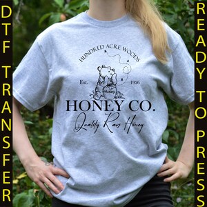 May include: A light gray t-shirt featuring a graphic of Winnie the Pooh with the text "Hundred Acre Woods" and "Honey Co. Quality Raw Honey." The design includes the year "1926" and the word "Hunny."