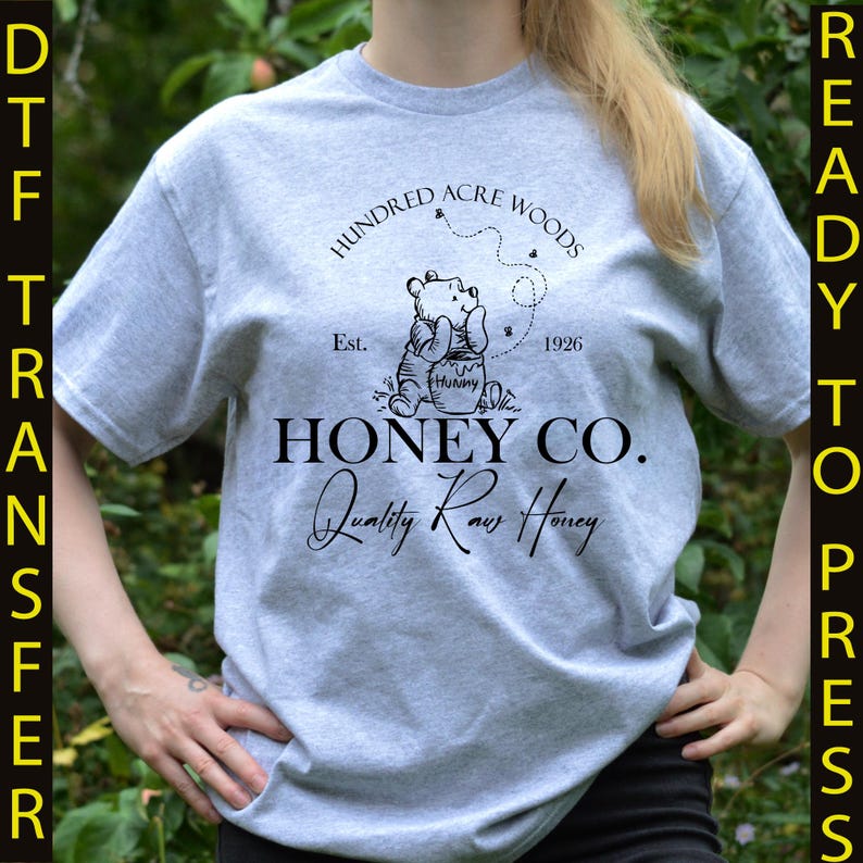 May include: A light gray t-shirt featuring a graphic of Winnie the Pooh with the text "Hundred Acre Woods" and "Honey Co. Quality Raw Honey." The design includes the year "1926" and the word "Hunny."