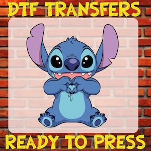 May include: A blue cartoon character with large black eyes and purple ears, forming a heart shape with its hands. The text "DTF TRANSFERS" and "READY TO PRESS" are in yellow, above and below the character, respectively.