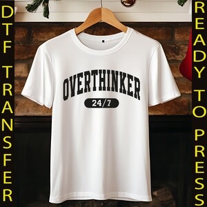 May include: White t-shirt with the text "OVERTHINKER 24/7" in black, arched lettering. The shirt is hanging on a wooden hanger against a brick fireplace background. The shirt is a DTF transfer, ready to press.