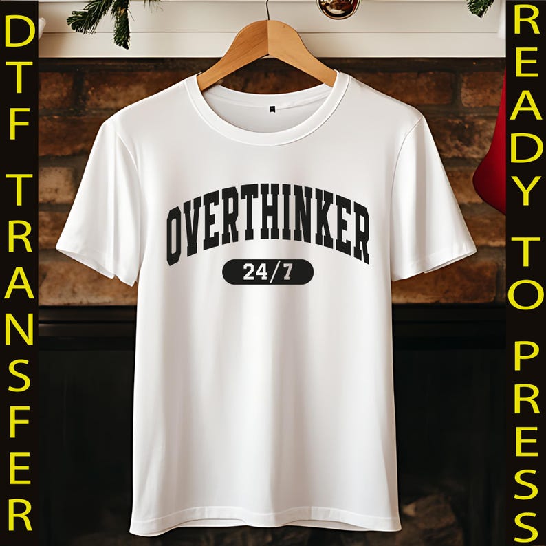 May include: White t-shirt with the text "OVERTHINKER 24/7" in black, arched lettering. The shirt is hanging on a wooden hanger against a brick fireplace background. The shirt is a DTF transfer, ready to press.