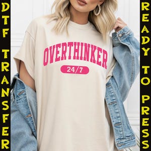 May include: A cream-colored t-shirt with the word "OVERTHINKER" in pink, arched across the chest, with "24/7" below. The shirt is worn with a denim jacket. The image has "DTF TRANSFER" and "READY TO PRESS" text on the sides.