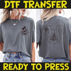May include: Gray t-shirt with a DTF transfer design. The front features a Mickey Mouse graphic with a castle and hearts. The back displays a castle design with the text "THE HAPPIEST PLACE ON EARTH" inside a Mickey Mouse head outline. The words "READY TO PRESS" are at the bottom.