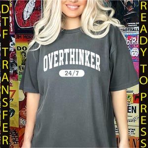 May include: Dark gray t-shirt with the text "OVERTHINKER 24/7" in white, arched across the chest. The shirt is a casual style, suitable for everyday wear. The background features colorful posters.