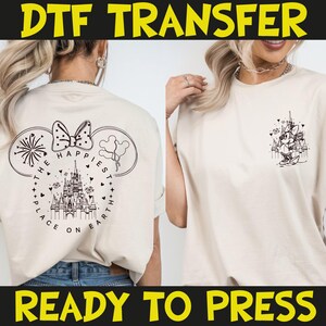 May include: Cream-colored t-shirt with a black graphic of a castle, fireworks, and Minnie Mouse ears. The text "THE HAPPIEST PLACE ON EARTH" is printed on the back. The words "DTF TRANSFER" and "READY TO PRESS" are printed in yellow.