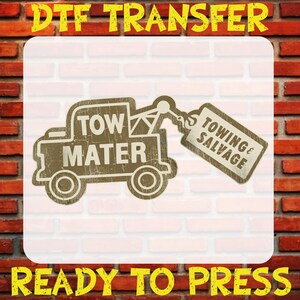 May include: A DTF transfer ready to press featuring a brown graphic of a tow truck with the text TOW MATER on the body and a hanging tag labeled TOWING & SALVAGE. The design has a distressed texture and is set against a white background.
