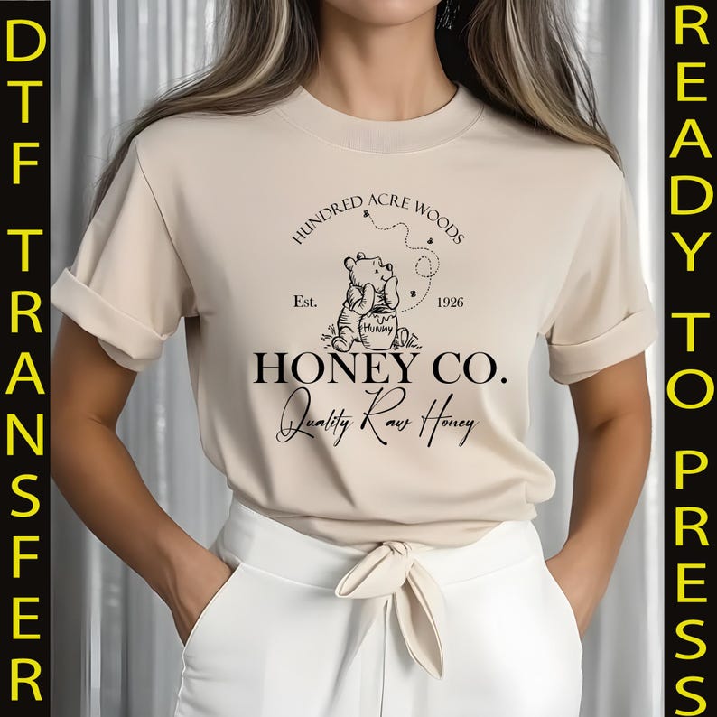 May include: Beige t-shirt featuring a graphic of Winnie the Pooh with the text "Hundred Acre Woods Honey Co. Quality Raw Honey." The design includes the year 1926. The shirt is a ready-to-press DTF transfer.