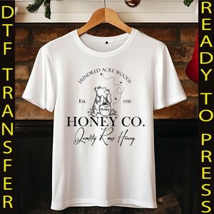 May include: White t-shirt featuring a graphic of Winnie the Pooh with the text "Hundred Acre Woods Honey Co. Quality Raw Honey." The design includes the year 1926 and the word "Hunny" on the honey pot.