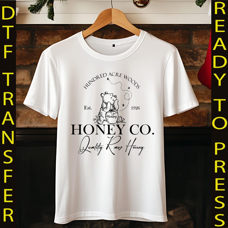 May include: White t-shirt featuring a graphic of Winnie the Pooh with the text "Hundred Acre Woods Honey Co. Quality Raw Honey." The design includes the year 1926 and the word "Hunny" on the honey pot.