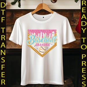May include: White t-shirt with a pink and blue baseball graphic. The graphic features a baseball diamond with a dripping pink icing design and the text "Baseball Grandma".