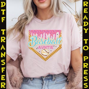 May include: Pink t-shirt with a baseball-themed graphic. The graphic features a waffle cone-shaped home plate with pink icing dripping down the sides. The text "Baseball Auntie" is written in blue and white lettering on the home plate. A baseball is in the center of the home plate.