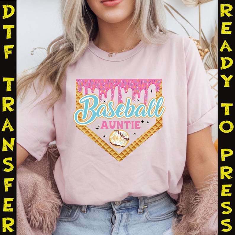May include: Pink t-shirt with a baseball-themed graphic. The graphic features a waffle cone-shaped home plate with pink icing dripping down the sides. The text "Baseball Auntie" is written in blue and white lettering on the home plate. A baseball is in the center of the home plate.