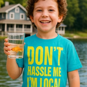 May include: A child wearing a turquoise t-shirt with the text "DON'T HASSLE ME I'M LOCAL" in yellow. The child is holding a glass of water with a goldfish. The background shows a lake and a house.