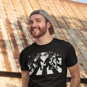 Puede incluir: Una camiseta negra con un gr&aacute;fico blanco de cuatro figuras caminando por un paso de cebra. Las figuras est&aacute;n estilizadas para parecerse a Bigfoot.