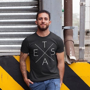 May include: A man wearing a black t-shirt with a white graphic of the word "TEXAS" in a crossed-out design. He is standing in front of a yellow and black striped wall.