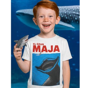 May include: A white t-shirt with the text "EL GRAN MAJA" in red and black, featuring a graphic of a large sea creature and a swimmer. A child is holding a toy shark. The background is a blue underwater scene.