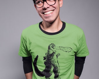 May include: A man wearing a green t-shirt with a black and white graphic of Godzilla breathing fire. He is smiling and looking to the right.