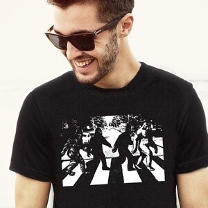 Puede incluir: Una camiseta negra con un gr&aacute;fico blanco de un grupo de figuras de Bigfoot caminando por un paso de cebra, similar a la portada del &aacute;lbum Abbey Road de los Beatles.