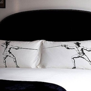 May include: Two white pillowcases with black illustrations of a fencer in action. The pillows are on a bed with a white comforter and a black headboard. A black nightstand with a lamp is to the right.