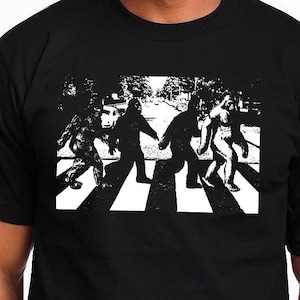 Puede incluir: Una camiseta negra con un gr&aacute;fico blanco de cuatro figuras de Pie Grande caminando por un paso de cebra, similar a la ic&oacute;nica portada del &aacute;lbum de los Beatles.