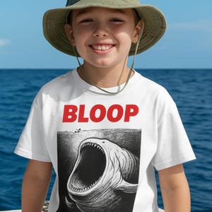 May include: White t-shirt with the word "BLOOP" in red letters. The shirt features a black and white illustration of a large fish with an open mouth. The person in the image is wearing a green hat.