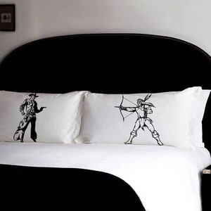 May include: Two white pillowcases with black embroidered images of a cowboy holding a gun and an archer with a bow and arrow. The pillows are on a black headboard with a white bedspread.