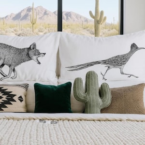 May include: White pillowcases with black and white animal illustrations of a fox and roadrunner. Decorative pillows include a green cactus, a dark green square pillow, a tan pillow, and a black and white patterned pillow. The bed has a white knitted blanket.