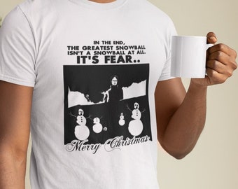 Könnte beinhalten: Ein weißes T-Shirt mit einer schwarzen Grafik einer Person, die auf einem schneebedeckten Hügel mit Schneemännern steht. Der Text auf dem Shirt lautet "In the end, the greatest snowball isn't a snowball at all. It's fear... Merry Christmas".