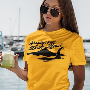 Może przedstawiać: Kobieta w ż&oacute;łtej koszulce z czarnym nadrukiem "Greetings from Loch Ness!" i czarną sylwetką potwora z Loch Ness. Trzyma zielony smoothie w szklanym słoiku ze słomką.