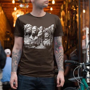 Puede incluir: Una camiseta marr&oacute;n con un gr&aacute;fico blanco que representa varias criaturas, incluyendo un gorila y una polilla. La camiseta la lleva una persona con tatuajes en los brazos. El fondo muestra un edificio de ladrillo y una bicicleta.