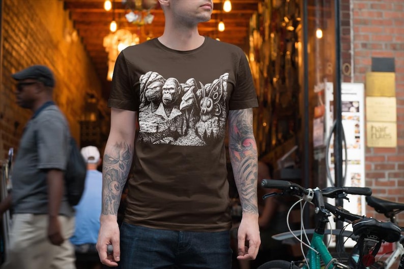 Puede incluir: Una camiseta marr&oacute;n con un gr&aacute;fico blanco que representa varias criaturas, incluyendo un gorila y una polilla. La camiseta la lleva una persona con tatuajes en los brazos. El fondo muestra un edificio de ladrillo y una bicicleta.