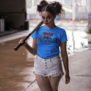 May include: A woman wearing a blue t-shirt with the text "Beware the Mothman!" and a black bat graphic. She is holding a baseball bat and wearing white denim shorts and black knee-high socks.