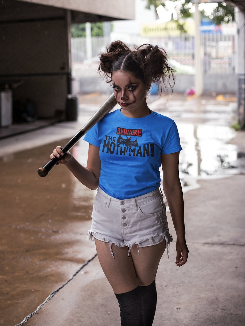 May include: A woman wearing a blue t-shirt with the text "Beware the Mothman!" and a black bat graphic. She is holding a baseball bat and wearing white denim shorts and black knee-high socks.