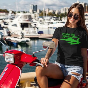 Może przedstawiać: Kobieta w czarnym t-shircie z zielonym nadrukiem potwora z Loch Ness i tekstem "Greetings from Loch Ness!" Siedzi na czerwonym skuterze w marinie.