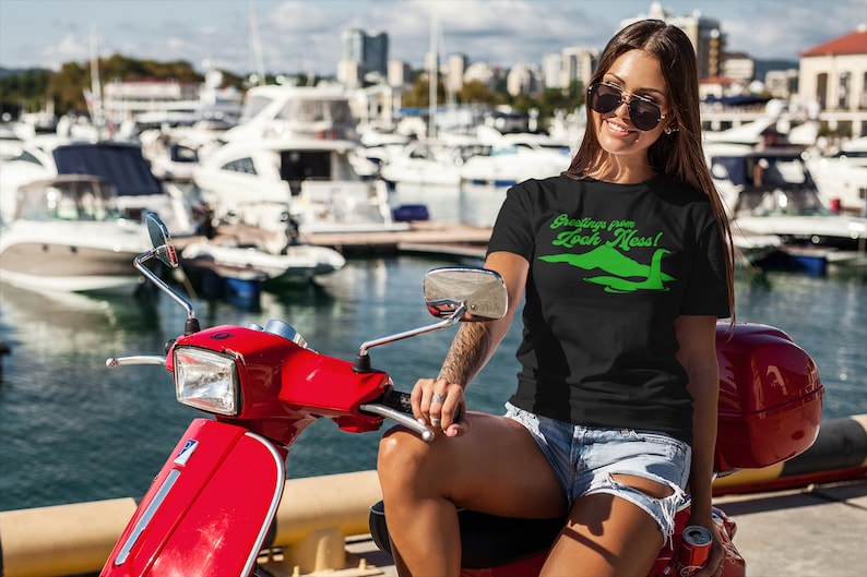 Może przedstawiać: Kobieta w czarnym t-shircie z zielonym nadrukiem potwora z Loch Ness i tekstem "Greetings from Loch Ness!" Siedzi na czerwonym skuterze w marinie.