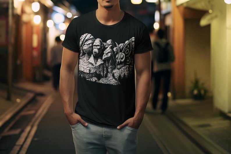 Puede incluir: Camiseta negra con un gr&aacute;fico blanco que representa un monumento con tem&aacute;tica de primates. El dise&ntilde;o incluye caras de gorilas y otros animales talladas en una estructura monta&ntilde;osa. La camiseta la lleva una persona con vaqueros azul claro.