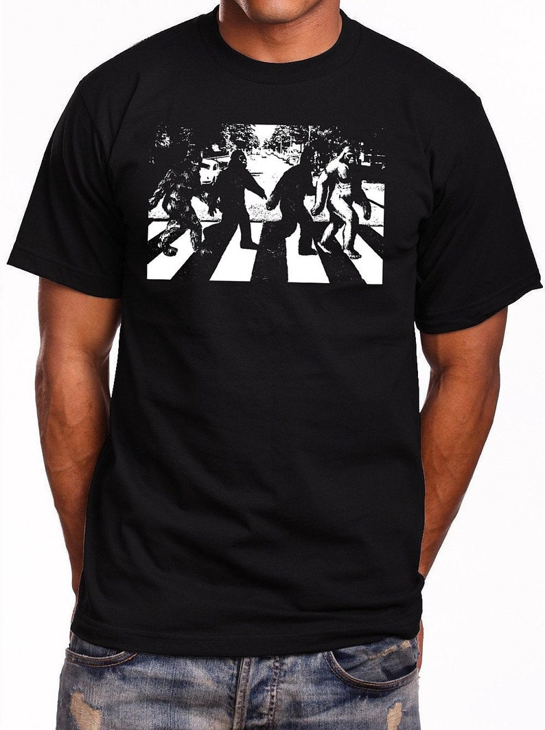 Puede incluir: Una camiseta negra con un gr&aacute;fico blanco de cuatro figuras de Pie Grande caminando por un paso de cebra, similar a la ic&oacute;nica portada del &aacute;lbum de los Beatles.