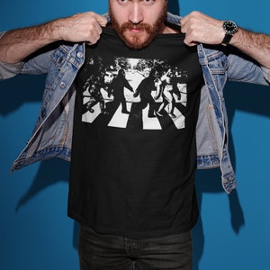 Puede incluir: Una camiseta negra con un gr&aacute;fico blanco de cuatro figuras de Pie Grande caminando por un paso de cebra, similar a la ic&oacute;nica portada del &aacute;lbum de los Beatles.